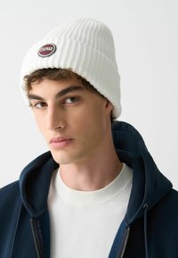 Biela rebrovaná čiapka s okrúhlym logom Colmar, nosená nad námorníckou mikinou a bielym sveterom s textúrovaným výstrihom.