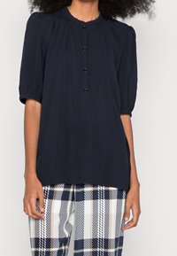 Blusa blu navy a maniche corte con scollo rotondo, chiusura a bottoni davanti e maniche a sbuffo. Il tessuto sembra leggero e morbido.