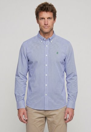 Hombre con camisa de cuadros azul claro abotonada y pantalones beige, de pie frente a un fondo liso gris claro.