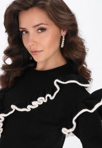 Maglione nero con dettagli di pizzo bianco lungo le maniche. La modella ha i capelli ricci e indossa orecchini a goccia di perle. Texture liscia, design aderente.