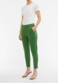 Pantalon vert sur mesure avec une taille ajustée, des plis à l'avant et des poches latérales. Associé à un haut blanc à manches courtes et des sandales transparentes.