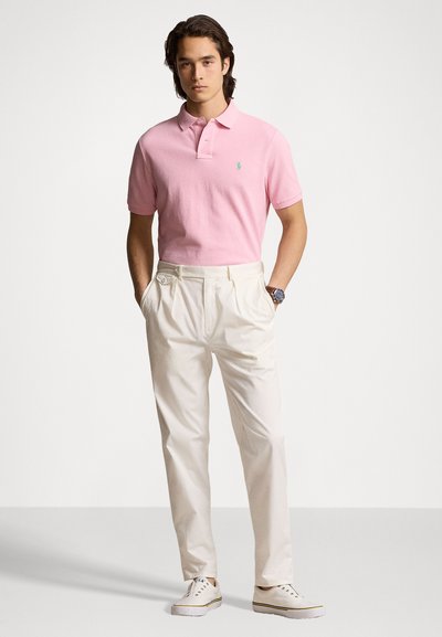 Cămașă polo roz cu mâneci scurte, logo mic pe piept, asortată cu pantaloni albi plisati și adidași albi. Croială casual, textură netedă.