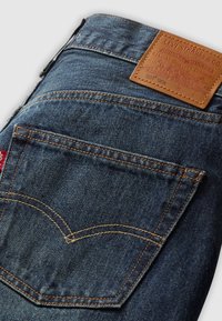 Poche de jean en denim bleu avec surpiqûres orange, étiquette en cuir Levi Strauss & Co., et étiquette en tissu rouge sur la couture latérale.