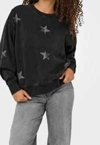 Sweat-shirt noir avec motifs d'étoiles en strass ; comporte un col rond et une coupe ample. Associé à un jean gris clair.