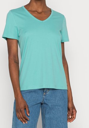 Kobieta ubrana w turkusowy T-shirt z krótkim rękawem i dekoltem w kształcie litery V oraz niebieskie dżinsy, stojąca z ramionami swobodnie opuszczonymi wzdłuż ciała.