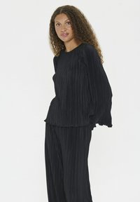 Donna con capelli ricci che indossa un top a maniche lunghe plissettato nero e pantaloni larghi plissettati abbinati, in piedi contro uno sfondo bianco.