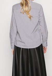 Valkoinen ja sininen raidallinen napillinen paita, jossa on pitkät hihat, kaareva helma ja takana yoke-yksityiskohta, yhdistetty mustaan nahkahameeseen.