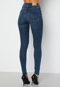 Štíhlé modré džíny vyrobené z denimu, s vysokým pasem, dvěma zadními kapsami a hladkou texturou. Nosené s černými lodičkami na vysokém podpatku.