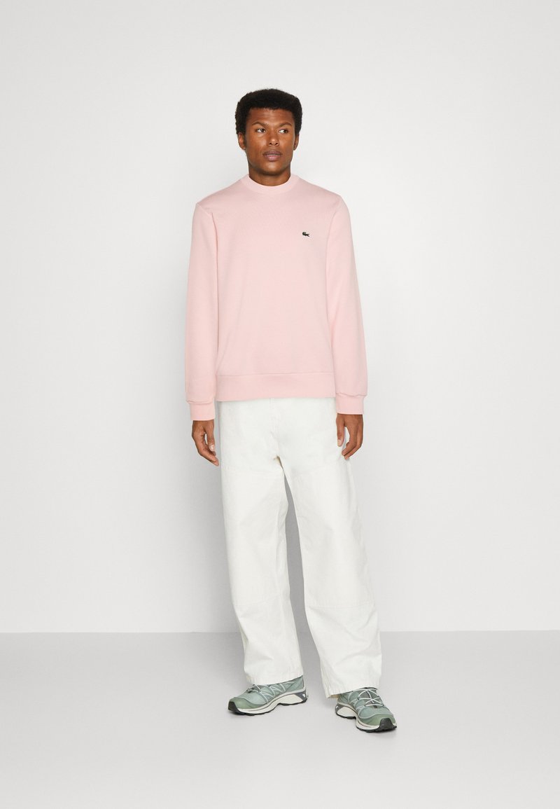 Sweatshirt rose clair avec un col ras du cou et des poignets côtelés, associé à un pantalon large blanc et des chaussures de sport grises. Détail de logo minimaliste.