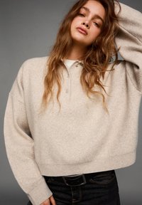 Világos bézs, rövidített pulóver, puha textúrával, gombos gallérral és ejtett vállakkal, sötét farmernadrággal és fekete övvel párosítva.