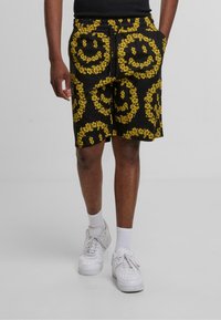 Svart shorts med gult blomstermotiv av smilende ansikt, elastisk midje, snor og sidelommer. Bært med hvite sneakers og sokker.