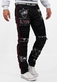 Pantaloni di jeans neri con strisce rosse sui lati, abbelliti con borchie e testo dipinto. Presentano una texture a coste e tasche con zip.