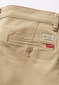 Pantalon en coton beige avec une poche arrière à fermeture par bouton, une étiquette portant la mention "Levi's" et des coutures distinctives le long des bords.