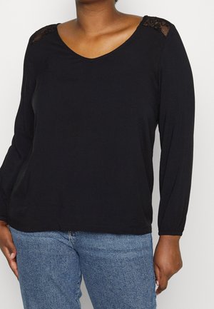 Kobieta ubrana w czarną bluzkę z długim rękawem z koronkowymi detalami na ramionach oraz jasnoniebieskie dżinsy, stojąca na jednolitym tle.