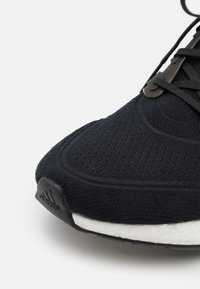 Zapatilla deportiva negra con parte superior de punto texturizada, que presenta una mediasuela blanca y una suela exterior de goma negra. Logo de Adidas notable en el talón.