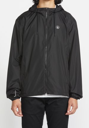 Person wearing a black hooded zip-up jacket with drawstrings, logo on chest, and elastic cuffs, paired with black pants and a white shirt underneath.