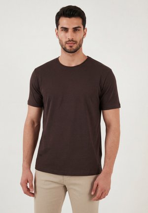 Ein Mann trägt ein dunkelbraunes T-Shirt mit kurzen Ärmeln und helle beigefarbene Hosen und steht vor einem einfarbigen hellen Hintergrund.