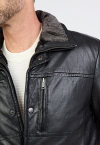 Man in een zwart leren jack met een ritszak op de borst en een grijze fleece-voering aan de kraag over een witte trui.