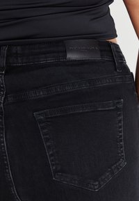 Svart denimjeans med en bakficka, en slät yta och en läderetikett vid midjan där varumärket är präglat.