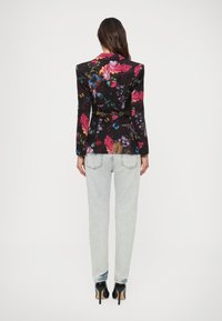 Donna in piedi con la schiena rivolta, indossa un blazer floreale nero, jeans azzurri chiari e scarpe con tacco nere su uno sfondo bianco.