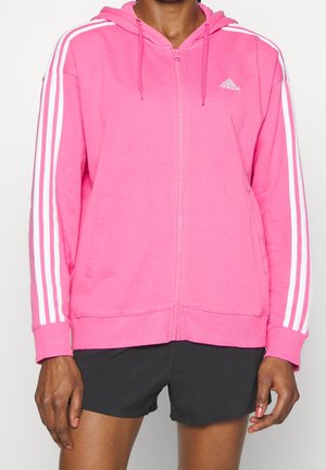 Osoba ubrana w jaskraworóżową bluzę z kapturem na zamek Adidas z białymi paskami na rękawach i czarne spodenki, pokazana od szyi do połowy uda.