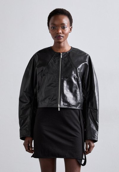 Chaqueta de cuero negra cortada con un acabado brillante, con cremallera frontal, mangas raglán y un diseño minimalista. Llevada sobre un vestido negro.