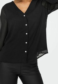 Blouse noire boutonnée en tissu transparent, avec un col en V et six boutons blancs sur le devant. Coupe ample mettant en valeur la silhouette.