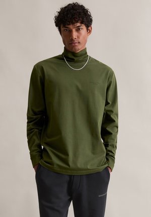 Grünes Langarm-Turtleneck-Shirt mit glatter Textur, das ein dezentes Logo auf der Brust trägt, kombiniert mit dunklen Hosen und einer silbernen Kette.