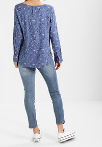 Blusa blu a maniche lunghe con motivo floreale in rosa e nero, orlo arrotondato e dettaglio con bottone sul retro, abbinata a jeans aderenti e scarpe da ginnastica bianche.