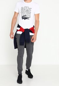 Vit t-shirt med svart texten "GAME TIME!" och krokodil-logotyp, kombinerad med grå sweatpants och en mörk blå-röd sweatshirt knuten i midjan.