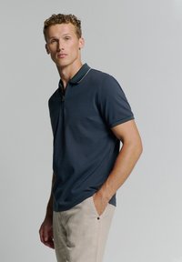 Marineblauwe polo met een ritssluiting bij de kraag, contrasterende afwerking, korte mouwen en een textuurstof. Gecombineerd met beige broeken met een knoopdetail.