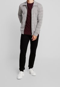 Mann in grauem karierte Jacke mit Reißverschluss, burgunderfarbenem Pullover, schwarzen hochgekrempelten Hosen und weißen Schnürschuhen, der vor einem grauen Hintergrund steht.