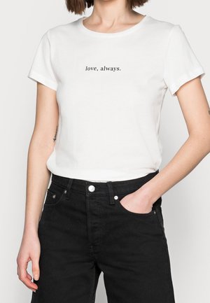 Kobieta w białej koszulce z napisem "miłość, na zawsze" i czarnych dżinsach, ręka w kieszeni, na jednolitym tle.