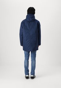 Marineblauwe hooded jack met een iets losse pasvorm, met een gladde textuur en een langere achterkant, gedragen over blauwe jeans en zwarte sneakers.