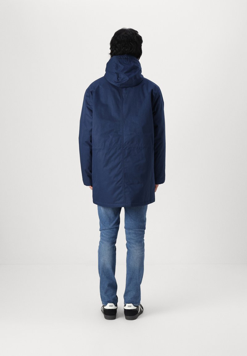 Giacca a vento blu navy con una vestibilità leggermente ampia, caratterizzata da una texture liscia e un retro più lungo, indossata sopra jeans blu e sneakers nere.
