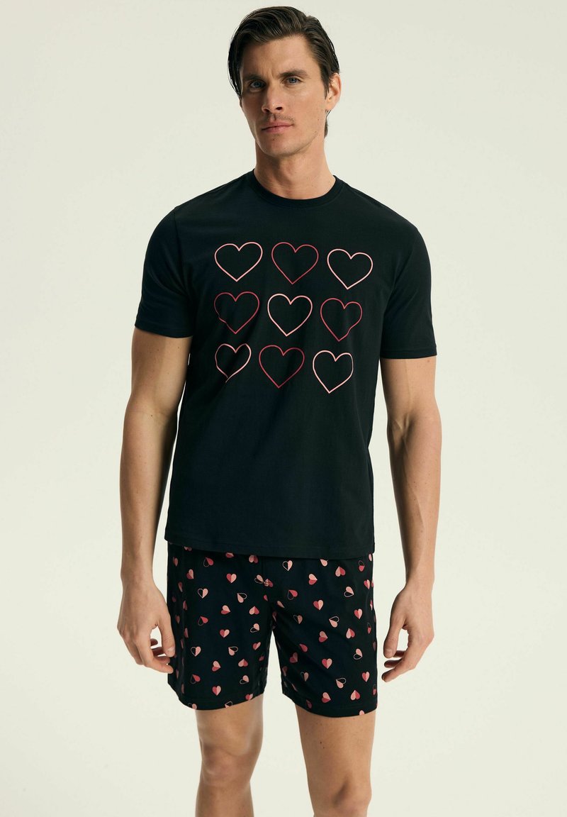 Czarna koszulka z wzorem serc w czerwonym konturze, zestawiona z czarnymi szortami z małymi, wielokolorowymi wzorami serc. Miękki materiał, codzienny krój.