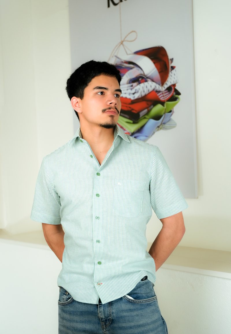 Jeune homme en chemise à manches courtes rayée vert clair et jeans, debout les mains derrière le dos, regardant vers sa gauche à l'intérieur près d'une œuvre d'art en tissus colorés pliés.