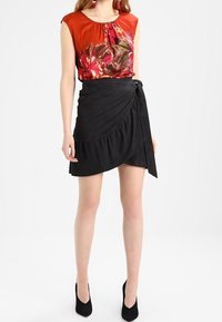 Kvinna i en röd blommig ärmlös topp instoppad i en högmidjad svart omlottkjol med svarta klackar.