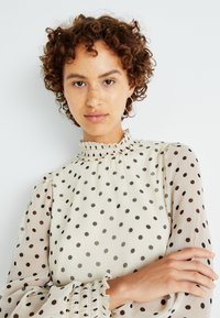 Šviesiai smėlinė palaidinė su juodais taškeliais, aukštas volaninis kaklas, permatomos pūstos rankovės ir elastingi rankogaliai. Lygi tekstūra, elegantiškas dizainas.