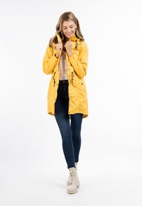 Gul vandtæt jakke med hætte, lynlåslukning, snor-detaljer og trykknapper, parret med mørke jeans og lyse støvler.