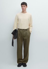 Camicia a maniche lunghe beige chiaro con texture, abbinata a pantaloni plissettati verde oliva. Modello tiene in mano una borsa in pelle nera. Design semplice e moderno.