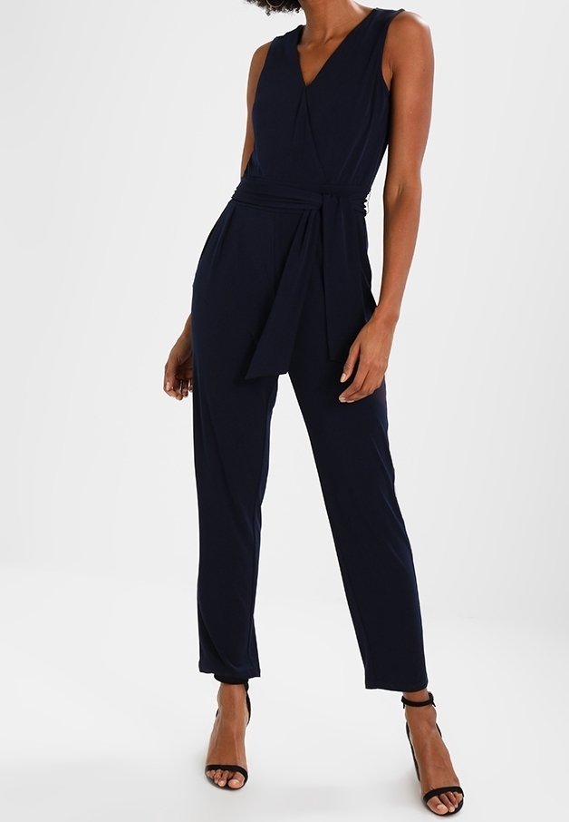 Femme portant une combinaison sans manches bleu marine avec col en V et ceinture en tissu, associée à des sandales à talons hauts noires avec bride à la cheville.