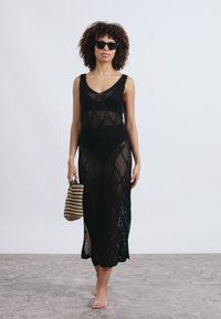 Μαύρο πλεκτό μάξι φόρεμα με υφή, που διαθέτει τετράγωνη λαιμόκοψη και πλαϊνές σχισμές, συνδυασμένο με τσάντα ριγέ και γυαλιά ηλίου.