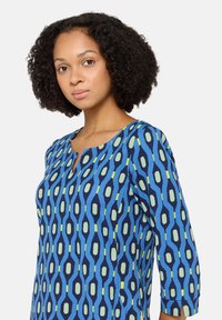 Blau gemustertes Kleid mit dreiviertel Ärmeln, das ein geometrisches Design in Grün und Gelb vor einem marineblauen Hintergrund sowie einen Schlüsselloch-Ausschnitt zeigt.