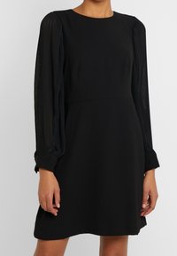Schwarzes Kleid mit strukturiertem Oberteil und langen, gefältelten transparenten Ärmeln. Das Design hat einen runden Halsausschnitt und reicht bis über das Knie.