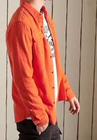 Oranssi flanellipaita, jossa napillinen etuosio ja kaksi rintataskua, valkoisen graafisen t-paidan päällä, esitetty neutraalilla taustalla.