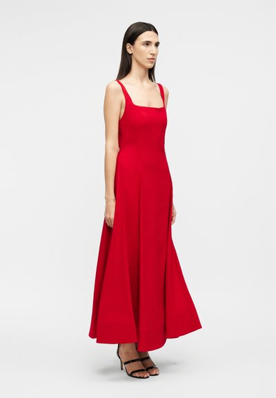 Vrouw met steil donker haar, gekleed in een mouwloze rode maxi-jurk en zwarte sandalen met hoge hak en bandjes, staand tegen een effen achtergrond.