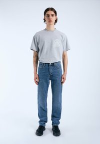 Camiseta de algodón gris con un diseño impreso en el lado izquierdo del pecho, combinada con vaqueros de mezclilla azul y zapatillas negras. Corte simple y relajado.