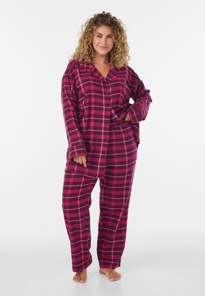 Curly-haired woman standing barefoot wearing loose, long-sleeve button-up and matching plaid pants in dark pink and magenta tones.