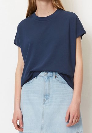 Femme portant un haut bleu marine à manches courtes, ample, rentré dans une jupe en denim bleu clair à boutons sur le devant et avec des poches.